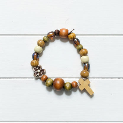 A beaded stretch bracelet featuring a mix of earthy brown, orange, olive green, and cream beads, a small white flower charm, and a light wood cross, displayed on a white background.