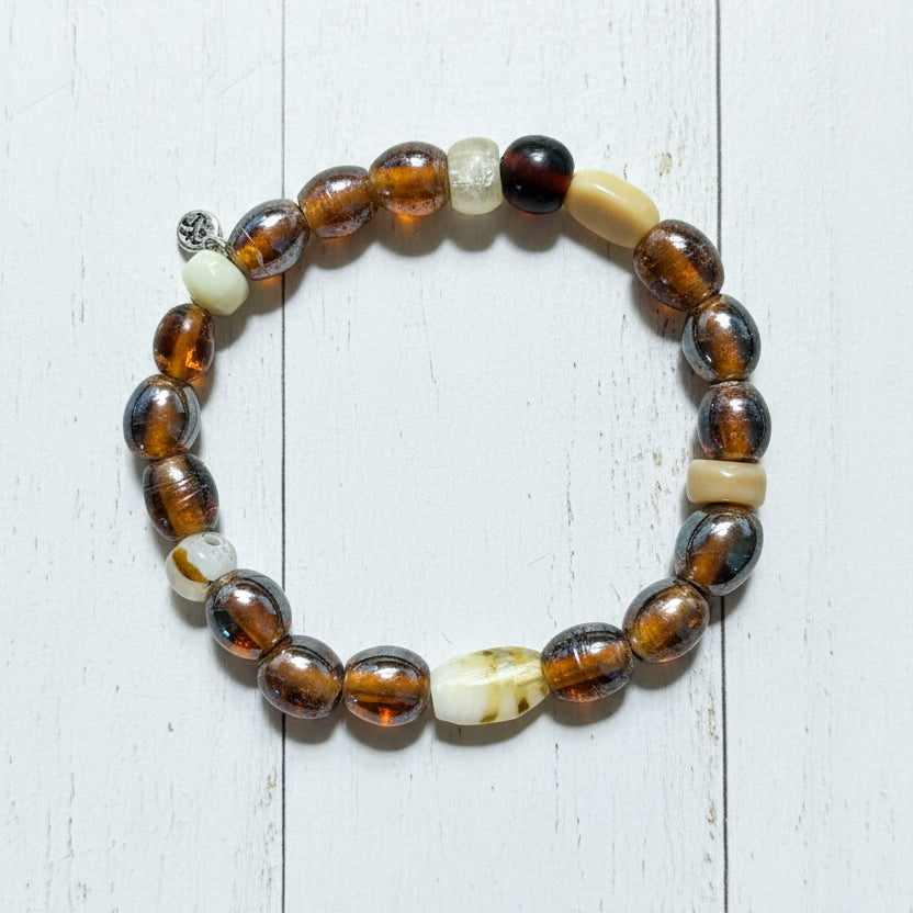 A beaded stretch bracelet featuring round brown and amber glass beads, polished tiger's eye-like beads, a white carved bead, a cream-colored cylindrical bead, and a prominent white carved accent bead, displayed on a white background.
