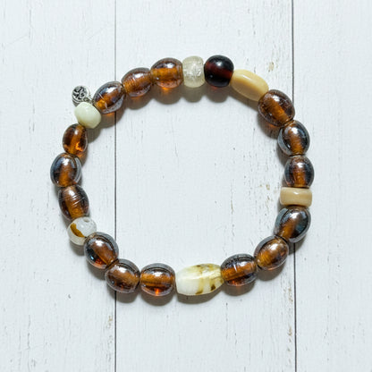 A beaded stretch bracelet featuring round brown and amber glass beads, polished tiger's eye-like beads, a white carved bead, a cream-colored cylindrical bead, and a prominent white carved accent bead, displayed on a white background.