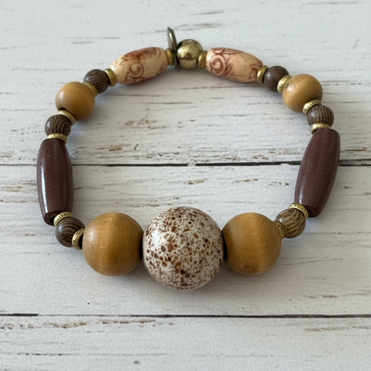 A beaded stretch bracelet featuring a central speckled brown and cream stone bead, flanked by two round gold-toned beads, a dark reddish-brown elongated bead, and alternating light wood and patterned beads on a white background.