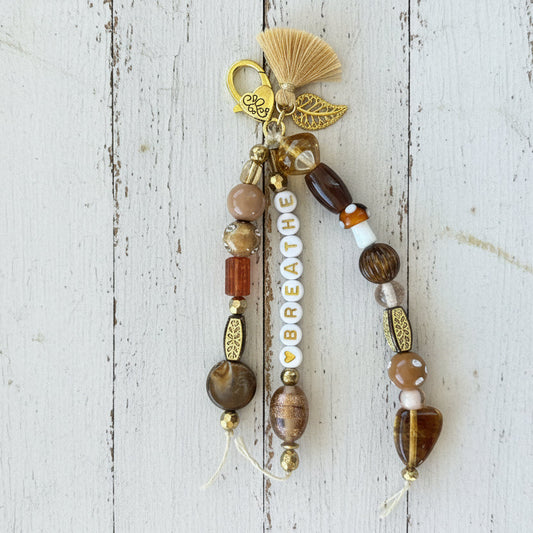 A handmade beaded keychain or phone strap with brown and gold beads, a gold angel charm, and white letter beads spelling 'BREATHE', laid on a white background.