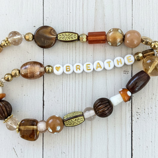 A close-up of a beaded bracelet laid flat on white background The bracelet features a mix of metallic gold-toned spacers, brown and amber glass beads, a white bead with a gold heart, and white letter beads spelling 'BREATHE'.