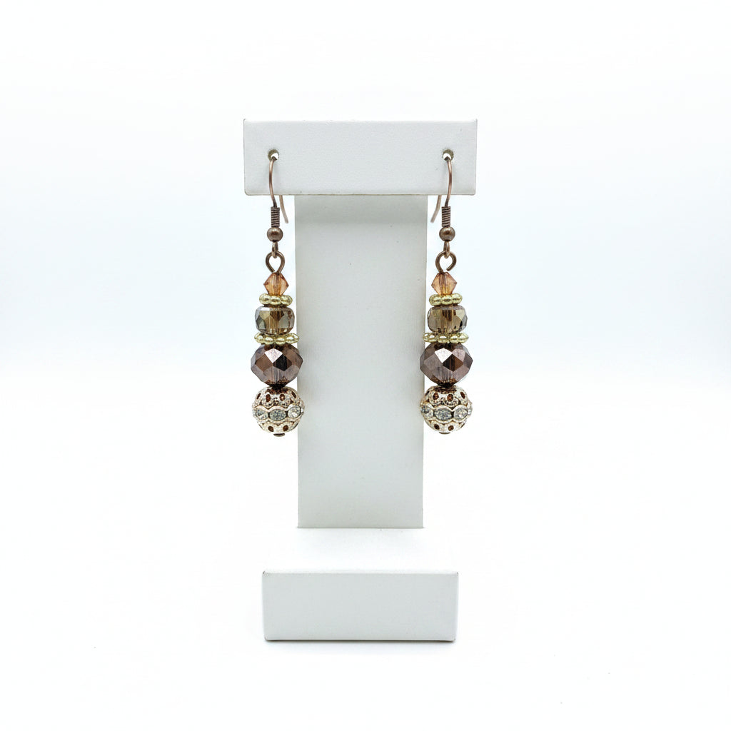 A pair of handmade dangle earrings with silver fishhook wires, faceted brown and iridescent beads, and a large ornate filigree ball at the bottom, displayed on a earring stand