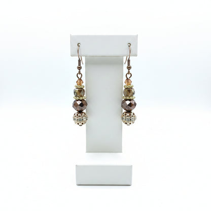 A pair of handmade dangle earrings with silver fishhook wires, faceted brown and iridescent beads, and a large ornate filigree ball at the bottom, displayed on a earring stand