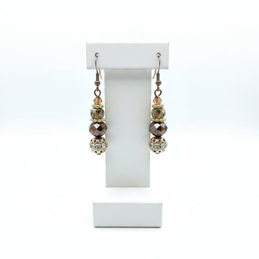A pair of handmade dangle earrings with silver fishhook wires, faceted brown and iridescent beads, and a large ornate filigree ball at the bottom, displayed on a earring stand
