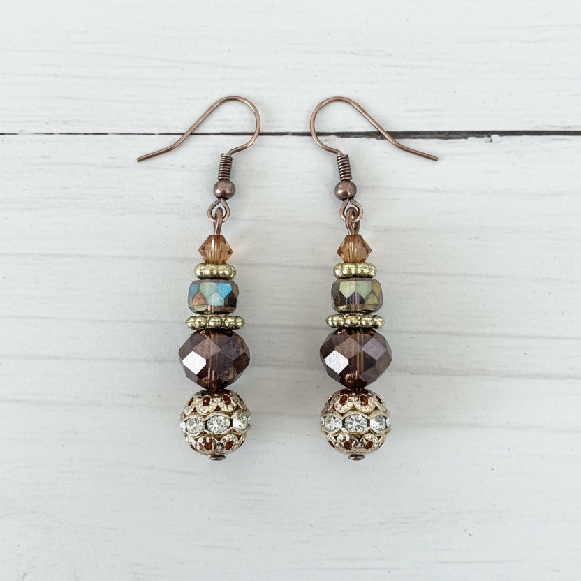 A pair of handmade dangle earrings with silver fishhook wires, faceted brown and iridescent beads, and a decorative gold-toned filigree ball at the bottom, displayed on a textured white surface.