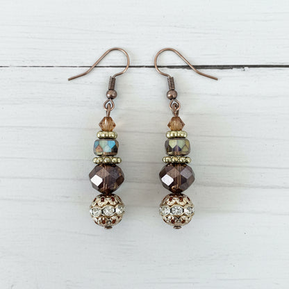 A pair of handmade dangle earrings with silver fishhook wires, faceted brown and iridescent beads, and a decorative gold-toned filigree ball at the bottom, displayed on a textured white surface.