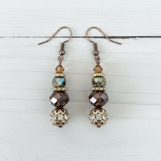 A pair of handmade dangle earrings with silver fishhook wires, faceted brown and iridescent beads, and a decorative gold-toned filigree ball at the bottom, displayed on a textured white surface.