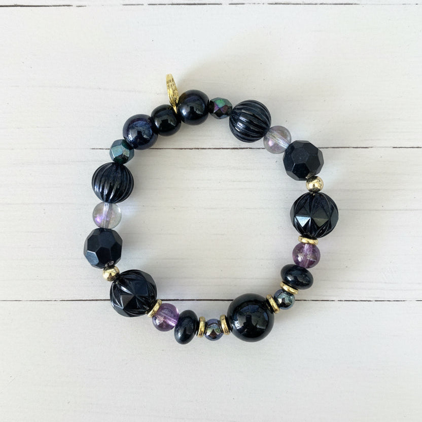 A beaded stretch bracelet featuring black faceted beads, dark blue iridescent beads, small purple beads, and gold spacer beads, displayed on a white textured paper background.