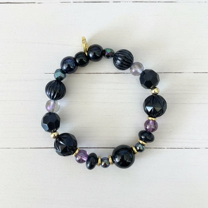 A beaded stretch bracelet featuring black faceted beads, dark blue iridescent beads, small purple beads, and gold spacer beads, displayed on a white textured paper background.
