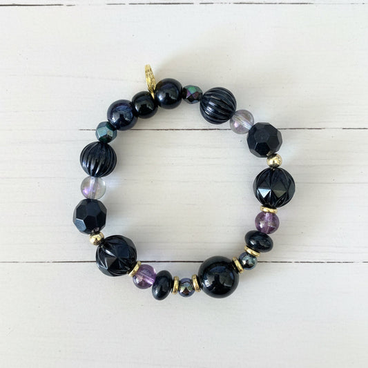 A beaded stretch bracelet featuring black faceted beads, dark blue iridescent beads, small purple beads, and gold spacer beads, displayed on a white textured paper background.