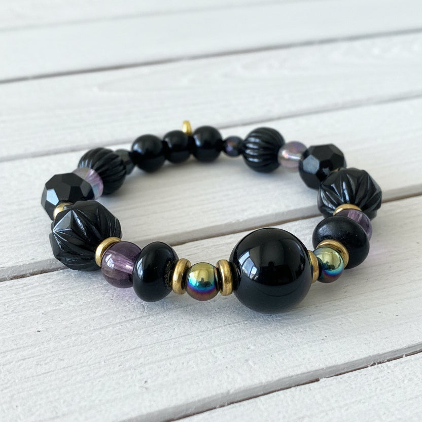 A beaded stretch bracelet featuring glossy black beads, faceted black beads, a purple bead, a metallic iridescent bead, and small gold spacer beads, displayed on a white textured surface.