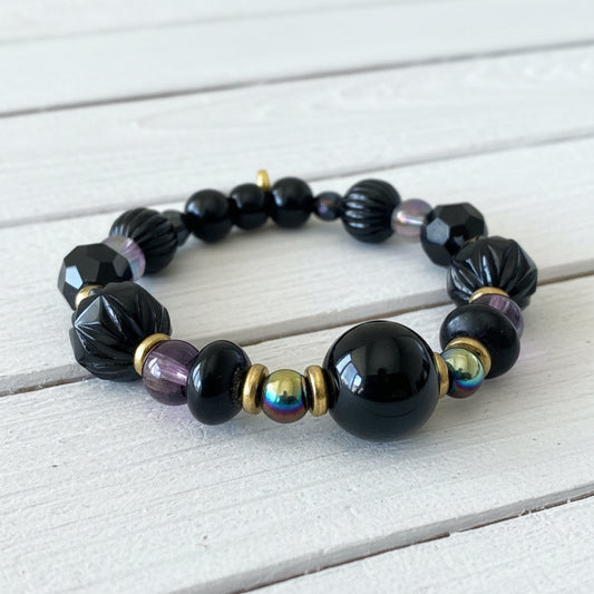 A beaded stretch bracelet featuring glossy black beads, faceted black beads, a purple bead, a metallic iridescent bead, and small gold spacer beads, displayed on a white textured surface.