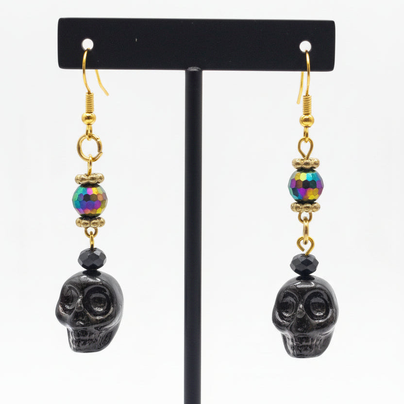 A pair of gothic-style drop earrings with gold-toned fishhook wires, iridescent metallic beads, and dark skull-shaped beads hanging on earring stand