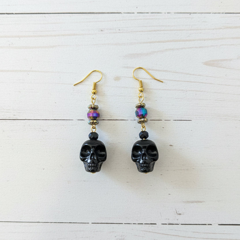 A pair of handmade dangle earrings with gold fishhook wires, small multicolored beads, and dark carved skull charms, resting on a white planks 
