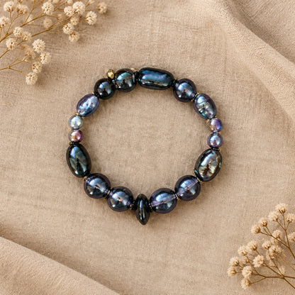 A beaded stretch bracelet featuring dark polished black and gray stones with small iridescent accents, displayed on a textured brown  background.