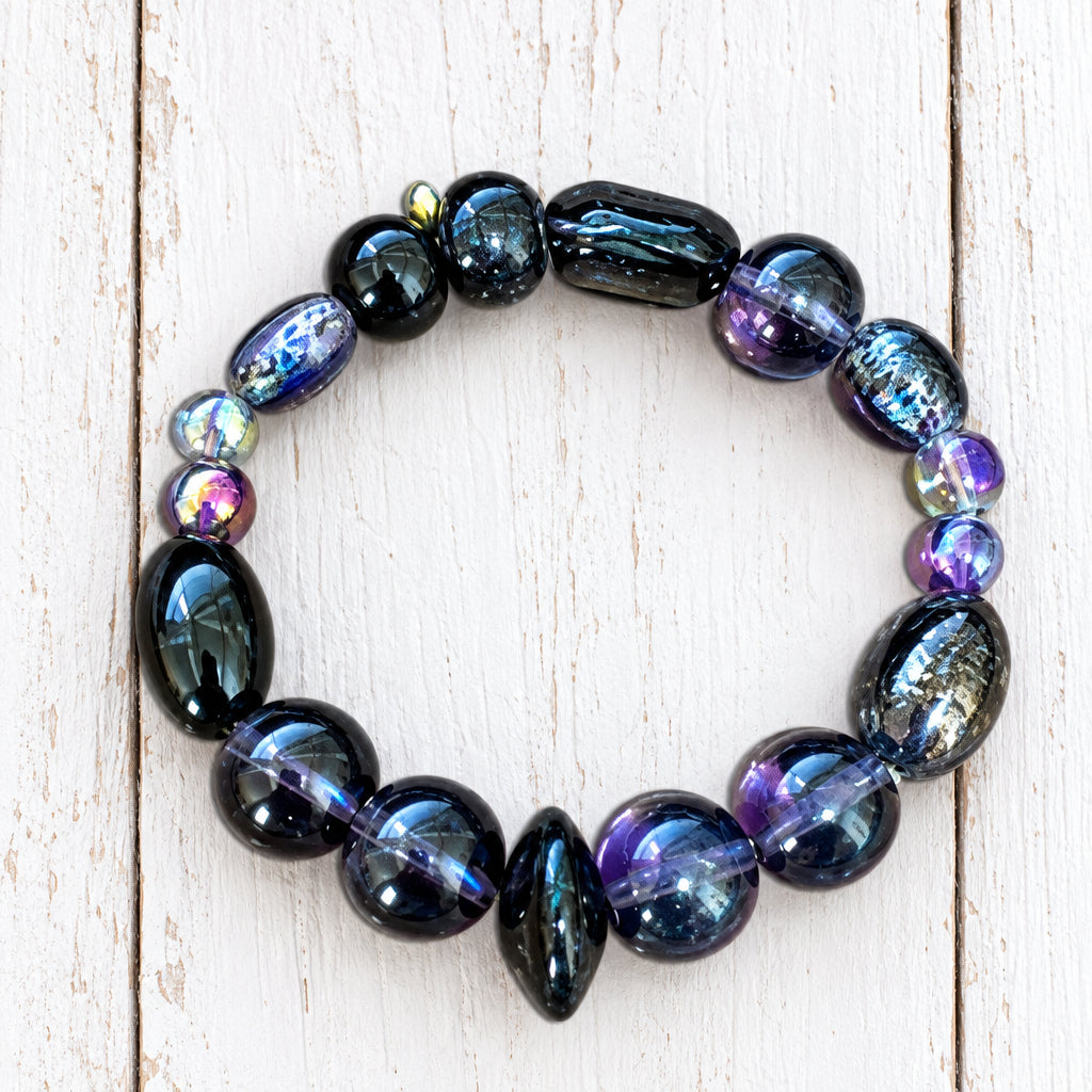 A beaded stretch bracelet made of glossy, dark iridescent beads with blue, purple, and black tones, displayed on a light background.