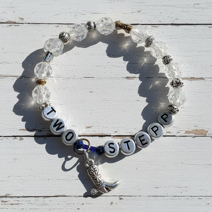 A beaded stretch bracelet with clear faceted beads, silver letter beads spelling 'TWO STEP', small silver spacer beads, a silver cowboy boot charm, and a dark blue cord accent, photographed on a light beige surface with strong shadows.