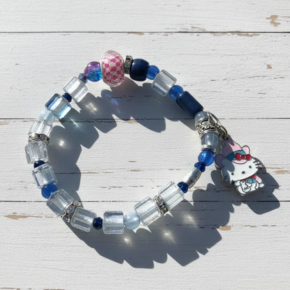 A beaded stretch bracelet featuring clear faceted beads, navy blue beads, a pink-and-white checkered bead, and a Hello Kitty charm, displayed on a beige background with soft shadows.